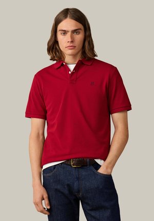 Ung mand med skulderlangt brunt hår iført en rød poloshirt og mørkeblå jeans med et flettet brunt bælte, stående foran en ensfarvet baggrund.