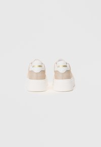 Sneaker platform beige con suole bianche spesse, linguette sul tallone bianche e branding dorato "Steve Madden", viste da dietro su uno sfondo semplice.