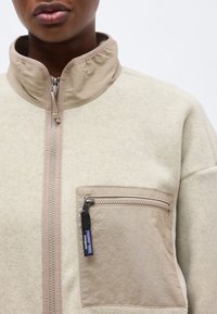 Κοντινό πλάνο ενός ατόμου που φοράει ένα μπεζ φλις μπουφάν με φερμουάρ μπροστά και τσέπη στο στήθος με φερμουάρ, που διαθέτει εικονίδιο της Patagonia ως τράβηγμα.