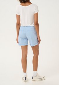 Kvinna stående med ryggen mot kameran, klädd i ljusblå uppvikta shorts, vit kortärmad tröja, vita strumpor och beigea sneakers.