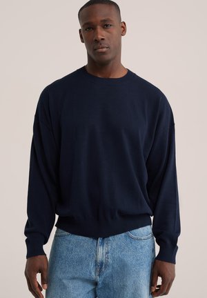 Mann trägt einen marineblauen Pullover mit langen Ärmeln und hellblaue Jeans, steht vor einem einfarbigen beigen Hintergrund.