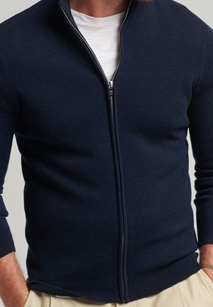 Mężczyzna w granatowym kardiganie na zamek błyskawiczny, na białej koszulce i beżowych spodniach, z rękami w kieszeniach, na neutralnym tle.