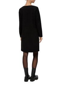 Robe noire à manches longues avec un décolleté rectangulaire, en tissu texturé avec des rayures horizontales, associée à des bottines noires à la cheville.