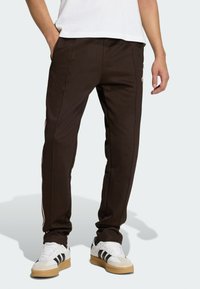 Hnedé teplákové nohavice s bočnými pruhmi, zúžený strih, elastický pás a vrecká. Skombinované s bielymi teniskami s čiernymi pruhmi.