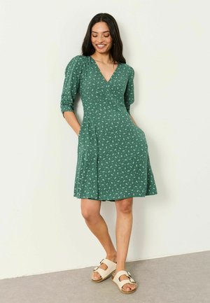Frau lächelt, trägt ein grünes knielanges Kleid mit weißem Muster, Dreiviertelärmel und beigen Sandalen, Hände in den Taschen, steht an einer Wand.