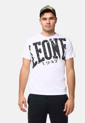Jeune homme portant un T-shirt blanc avec le texte "LEONE 1947", un pantalon noir et une casquette noire et jaune à motif, debout devant un fond uni.