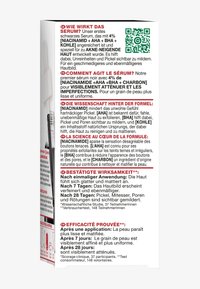 Svart serumförpackning med vit och röd text som lyfter fram aktiva ingredienser: Niacinamid, AHA, BHA och kol. QR-kod ingår.