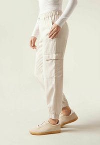 Beige cargo kalhoty s elastickými manžetami, bočními kapsami a uvolněným střihem, kombinované se světlými teniskami. Hladká textura, ležérní design.