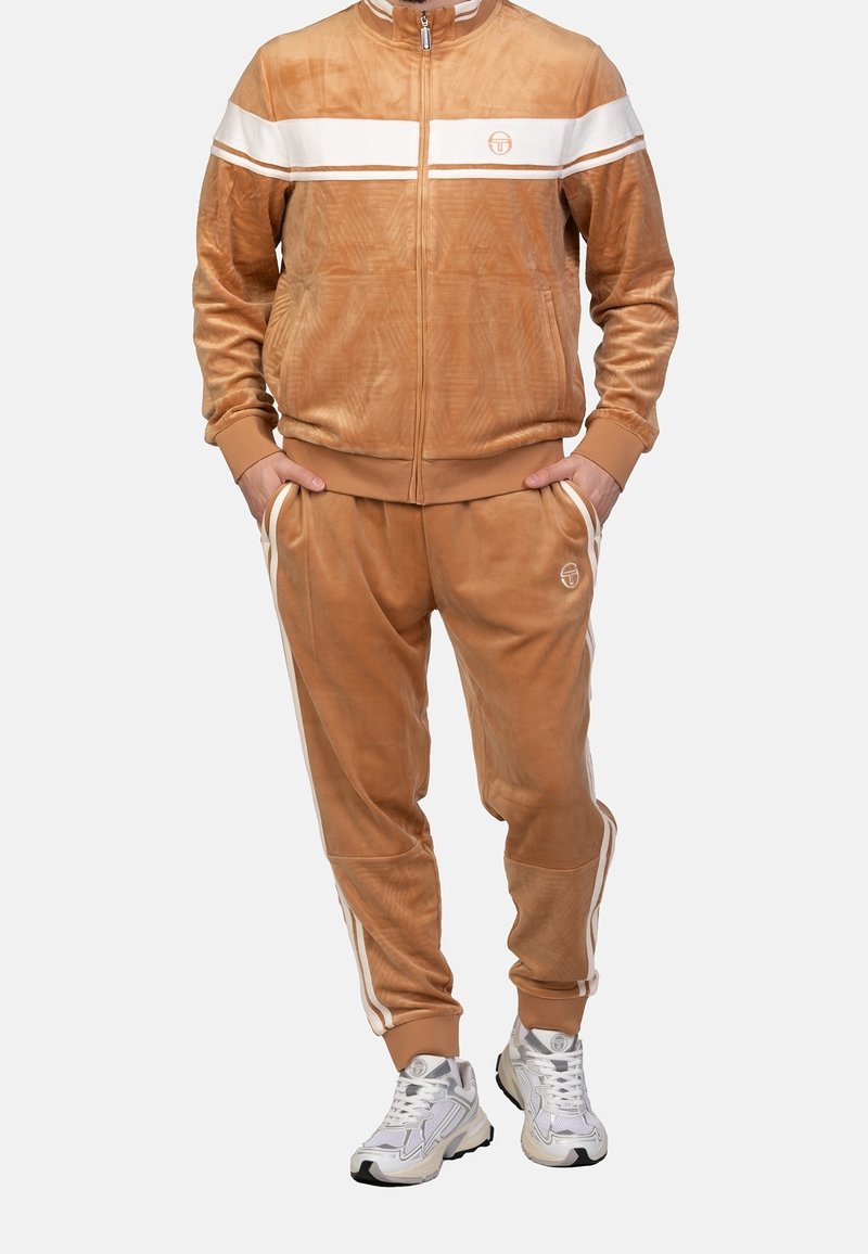 Homme portant un survêtement en velours marron camel avec des bandes blanches et un logo, assorti à des chaussures de sport blanches et grises, debout les mains dans les poches.