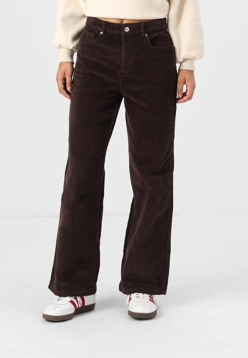Femme portant un pantalon large en velours côtelé marron foncé, un pull court crème et des baskets blanches avec des rayures rouges, debout devant un fond blanc.