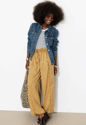Femme souriante aux cheveux naturels portant une veste en jean, un haut rayé, un pantalon large beige, des sandales noires et un sac à imprimé léopard.