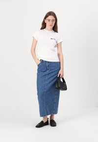 Witte t-shirt met een klein zwart logo, gecombineerd met een enkellange blauwe denim rok. Een zwarte handtas en zwarte loafers maken de outfit compleet.