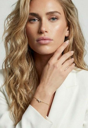 Lyshåret kvinde med bølget hår iført en hvid blazer og et delikat guldarmbånd med tre runde diamanter på håndleddet.