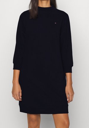 Femme portant une robe-pull noire unie à manches longues avec un petit logo Tommy Hilfiger sur la poitrine, debout devant un fond blanc.