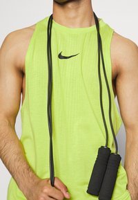 Hombre vistiendo una camiseta sin mangas de color verde lima de Nike, sosteniendo las empuñaduras de una cuerda para saltar negra frente a un fondo liso.