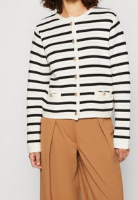 Cardigan a righe bianche e nere con bottoni dorati, texture a coste e due tasche frontali. Indossato sopra pantaloni marroni a vita alta.