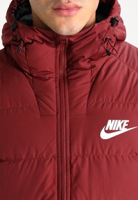 Marinröd puffjacka med hög krage, framzipper och svarta detaljer. Har en vit Nike-logotyp på bröstet. Strukturerad tyg.