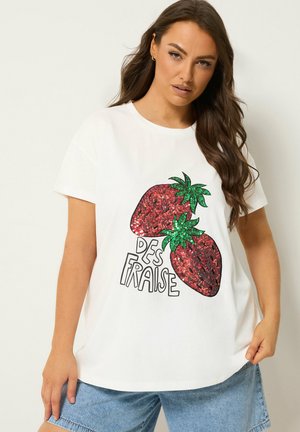 Mujer con una camiseta blanca con dos grandes fresas con lentejuelas y texto "Des Fraise", combinada con shorts vaqueros azul claro.