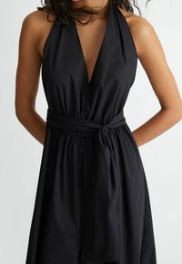 Schwarzes Halsband-Kleid mit tiefem V-Ausschnitt, tailliertem Bund und seitlichen Taschen. Hergestellt aus glattem Stoff mit fließender Silhouette.