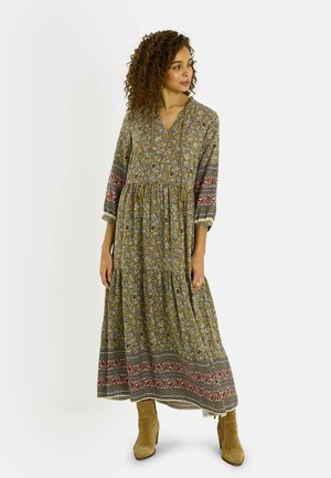 Mujer con cabello rizado que lleva un vestido largo maxi verde oliva con estampado floral en las mangas y el dobladillo, combinado con botines de ante color canela.