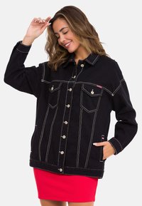 Schwarze Jeansjacke mit kontrastierenden weißen Nähten, Fronttaschen, Metallknöpfen und einem Kragen, kombiniert mit einem roten Rock.