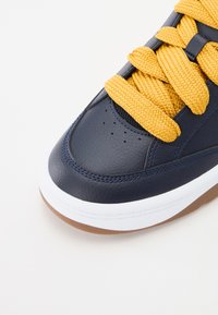 Ναυτικό δερμάτινο αθλητικό παπούτσι με υφή, κίτρινες υφαντές κορδόνες, λευκή ενδιάμεση σόλα και σόλα από καουτσούκ γόμας; διαθέτει τρυπήματα στο μπροστινό μέρος.