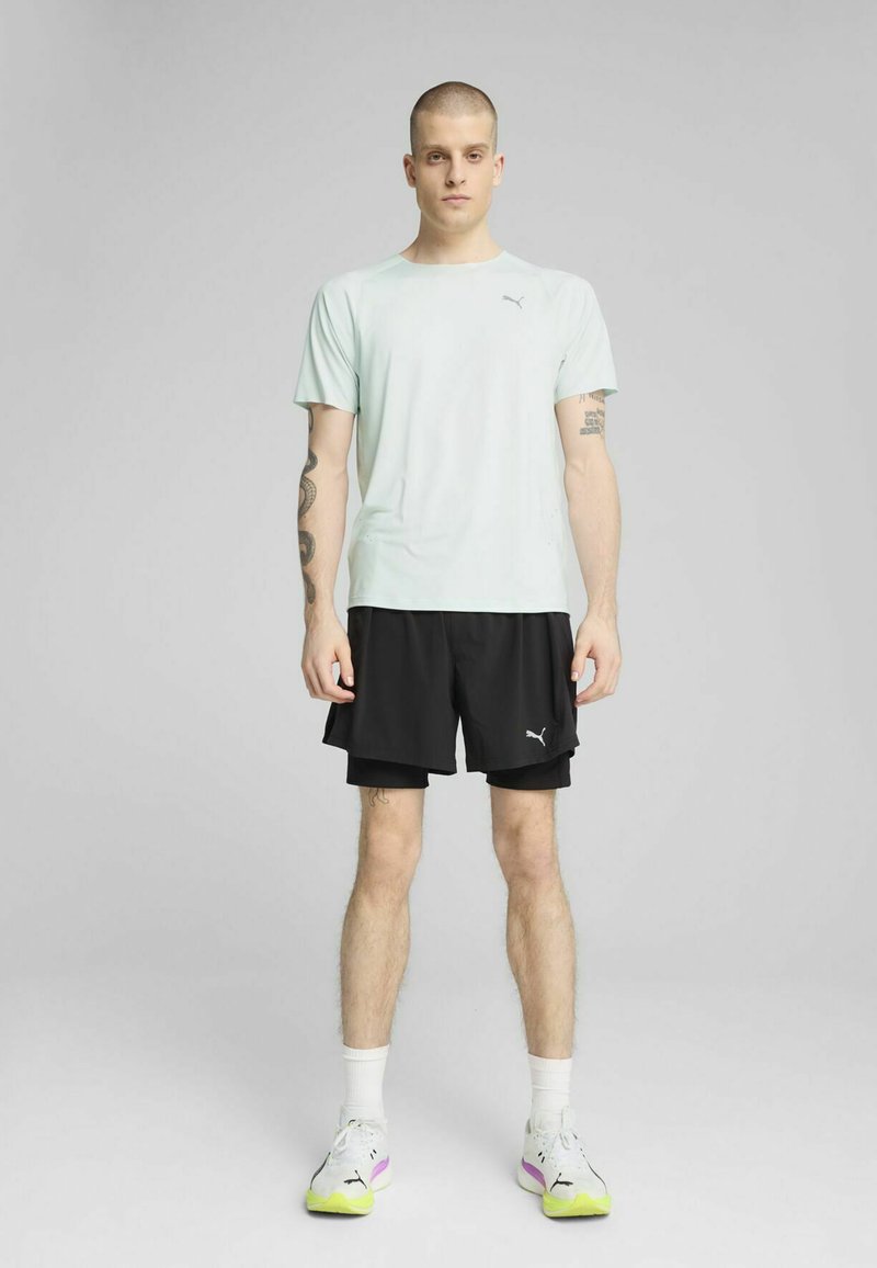 Lysgrøn kortærmet skjorte med et logo på brystet, parret med sorte shorts. Sættet er komplementeret af hvide sneakers med farverige accenter.