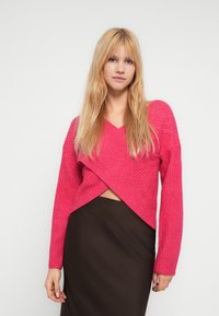 Fuksia ribbestrikket genser med V-hals og kryssdetalj, kombinert med et brunt skjørt, med lange ermer og en teksturert overflate.