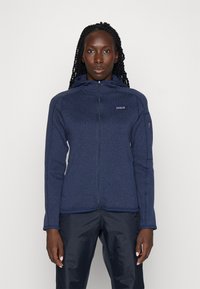 Marineblå hettejakke med glidelås laget av strukturert fleece, med høy krage, sidelommer og et logo-patch på brystet.