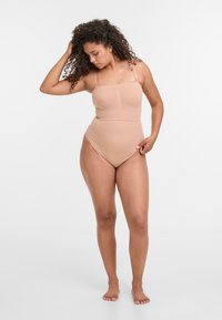 Beiger Body mit dünnen Trägern, figurbetontem Design, glatter Textur und hohen Beinausschnitten. Das Model posiert mit der Hand im Haar und zeigt die Passform.