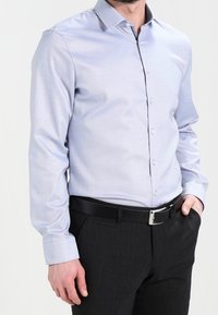 Ljusblå långärmad skjorta med knappkrage, tillverkad av slitstark tyg. Bärs tillsammans med svarta byxor och ett stilrent bälte.