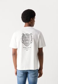 T-shirt bianco in cotone con una grafica di tigre in linea nera sul retro, con testo del marchio sottostante. Maniche corte e scollatura rotonda.