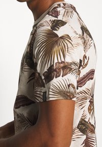 Beige T-shirt met korte mouwen, voorzien van een kleurrijk palmblaadjesprint in groen, bruin en bordeauxrood, met een ronde hals en subtiele stikdetails.