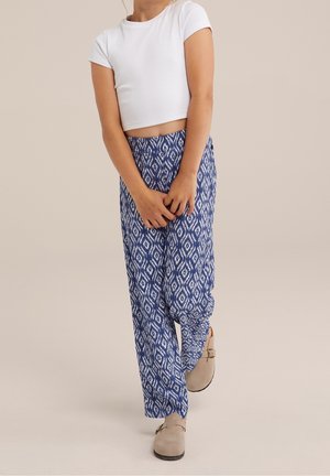 Personne portant un crop top blanc à manches courtes et un pantalon large bleu à motifs avec des chaussures beiges à enfiler, debout sur un fond uni.
