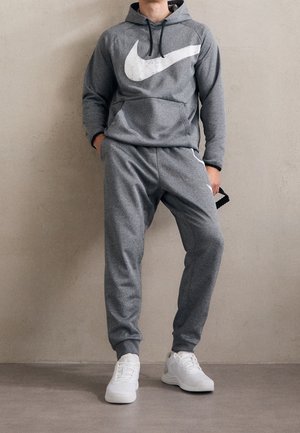 Sudadera gris con un gran logo blanco de Nike, combinada con pantalones deportivos a juego; incluye puños acanalados y zapatillas deportivas blancas.