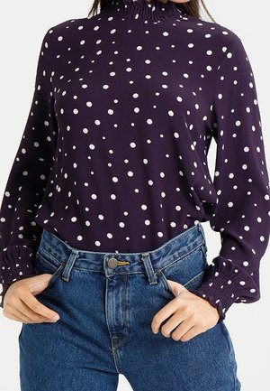 Frau trägt eine lila Langarmbluse mit weißen Punkten und hoch taillierte blaue Jeans, Hände auf den Hüften ruhend.