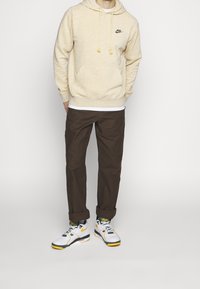 Nike hoodie in light tan with a front pocket and drawstrings, paired with dark brown pants and white/black sneakers with green accents.