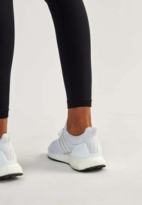 Baskets de sport blanches avec tige en mesh, design texturé et semelle en caoutchouc, associées à un legging noir, mettant en valeur une silhouette ajustée.