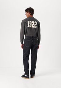 Camisa de algodón gris de manga larga con "1922" impreso en crema en la parte de atrás, combinada con jeans de mezclilla oscura y zapatos negros.