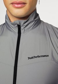 Hellgraue Zip-Jacke aus glattem Stoff mit Stehkragen und schwarzem Reißverschluss. "PeakPerformance"-Logo auf der Brust eingestickt.