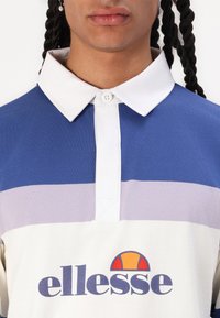 Polo de manga curta em azul, branco e riscas em roxo claro. Apresenta um colarinho branco, placket com botões e o logótipo "ellesse" na frente.