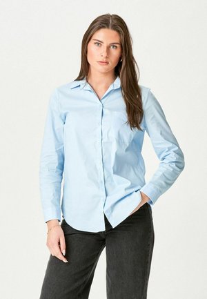 Junge Frau mit langem braunem Haar, die ein hellblaues Hemd mit Knöpfen und schwarze Hosen trägt, stehend mit einer Hand in der Tasche, neutrale Hintergrund.