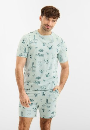 Mies, joka pukeutunut vaaleanvihreään, kuvioituun t-paitaan ja shortseihin, joissa on tummanvihreitä rantateemaisia kuvia, seisoo yhdellä kädellä taskussa.