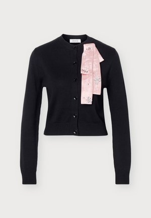 Un mannequin se tient devant un fond clair, vêtu d’un cardigan noir boutonné, d’une écharpe rose, d’une jupe sombre à fleurs et à franges, et de chaussures noires à talons.