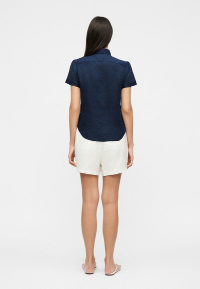 Femme debout, vue de dos, portant un t-shirt bleu marine à manches courtes, un short blanc et des sandales plates sur un fond uni.