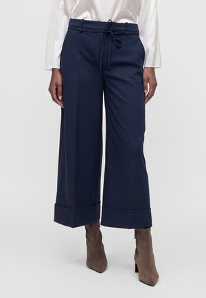 Femme portant un pantalon large bleu marine raccourci avec des ourlets retournés, une taille nouée, un chemisier blanc à manches longues et des bottines taupe à talons.