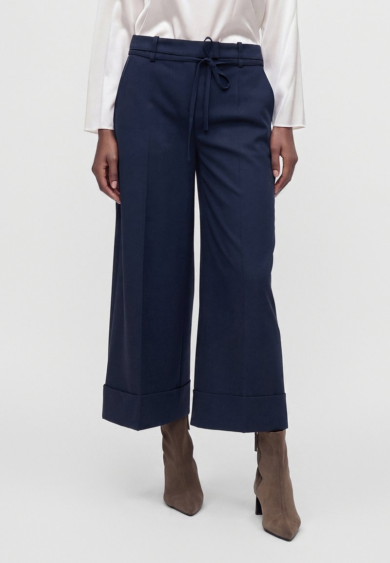 Femme portant un pantalon large bleu marine raccourci avec des ourlets retournés, une taille nouée, un chemisier blanc à manches longues et des bottines taupe à talons.