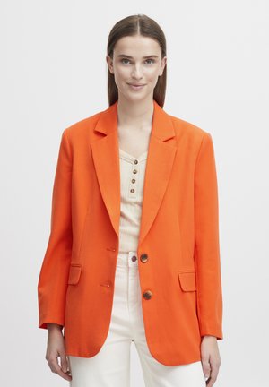 Orange blazer med klaplommer, enkelt knaplukning og to frontlommer. Stilet over en beige top og parret med hvide bukser.