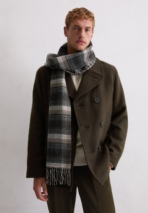 Wollen sjaal in grijstinten met een geruit patroon en gefranjerde randen, gedragen over een olijfgroene jas met dubbele knopen en een getextureerde kraag.