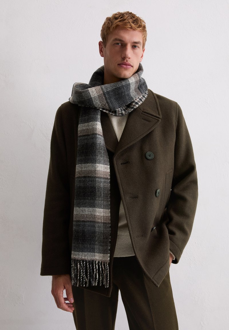 Wollen sjaal in grijstinten met een geruit patroon en gefranjerde randen, gedragen over een olijfgroene jas met dubbele knopen en een getextureerde kraag.